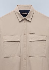 Camicia beige con bottoni e colletto classico, caratterizzata da due tasche frontali con pattina e un'etichetta con logo sotto il colletto, realizzata in tessuto liscio.