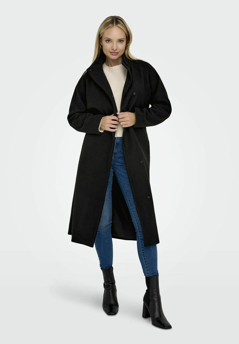 Manteau long noir en matériau doux avec un col, des boutons sur le devant et deux poches latérales, associé à un jean bleu et des bottines noires.