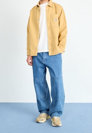 Chemise en coton jaune superposée à un t-shirt blanc, associée à un pantalon en denim bleu. Les chaussures sont beiges avec un design texturé.
