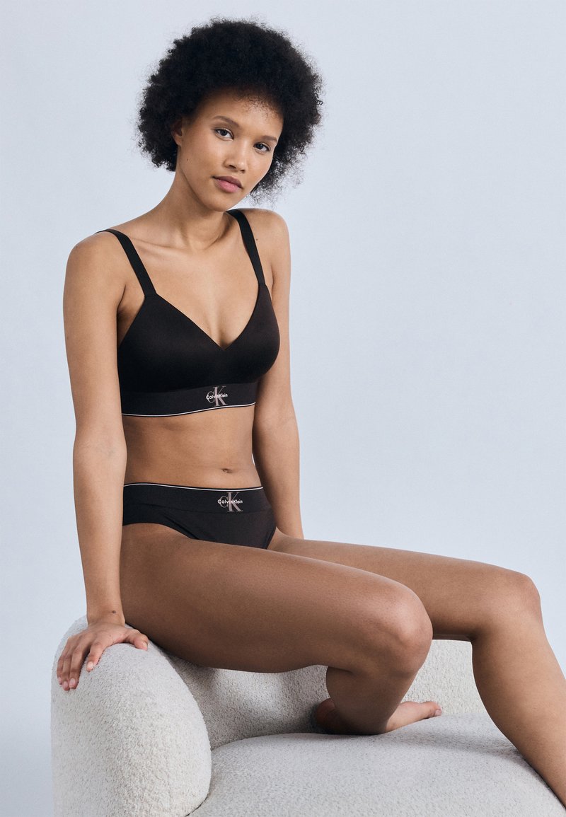 Γυναίκα με κοντά σγουρά μαλλιά που φοράει μαύρο σουτιέν και εσώρουχα Calvin Klein, καθισμένη σε μια υφασμάτινη πολυθρόνα ανοιχτού γκρι χρώματος σε απλό φόντο.