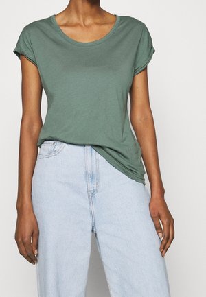 Person iført en grøn kortærmet T-shirt, let stoppet ind i lyseblå højhøjede jeans, stående mod en ensfarvet baggrund.