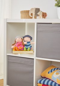 Zwei Stoffpuppen sitzen auf einem Stapel Bücher auf einem weißen Regal. Die Puppen tragen bunte Outfits. Unter ihnen befinden sich graue Stofflagerbehälter.