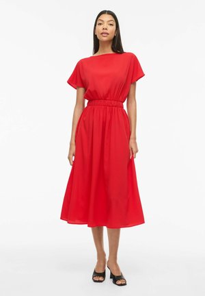 Kvinde stående iført en knaldrød midi-kjole med korte ærmer og talje, parret med sorte åbne peep-toe blokhæle sko mod en hvid baggrund.
