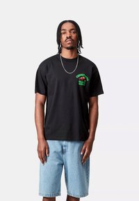 Camiseta de algodón negra con diseño gráfico verde que presenta texto y un motivo en el lado izquierdo del pecho. Combinada con pantalones cortos de mezclilla azul claro.