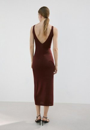 Kobieta stoi tyłem, ubrana w bordową sukienkę midi bez rękawów z głębokim dekoltem na plecach oraz czarne sandały na wysokim obcasie, na jednolitej szarej podłodze.