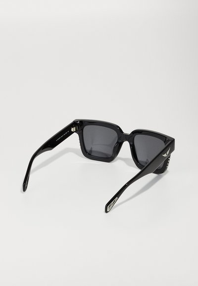 Occhiali da sole neri con lenti quadrate, montatura spessa e lucida, lenti scure, dettagli in metallo sulle aste e logo con finitura testurizzata.