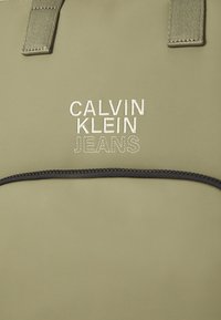 Olivová taška cez rameno s čiernym zipsom a rúčkami. Na prednej strane je vyšívaný nápis "CALVIN KLEIN JEANS" v svetlosivej farbe tučným písmom.