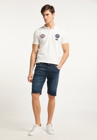 Camiseta polo blanca con parches bordados, combinada con pantalones cortos de mezclilla oscuros y zapatillas blancas. El ajuste es casual, con un collar estándar y mangas cortas.