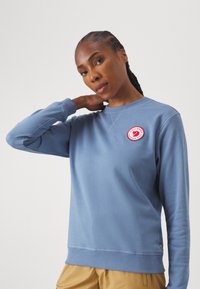 Hellblaues Sweatshirt mit Rundhalsausschnitt, gerippten Bündchen und Saum. Verfügt über ein rundes rotes Patch-Logo auf der linken Brust und strukturierten Stoff.
