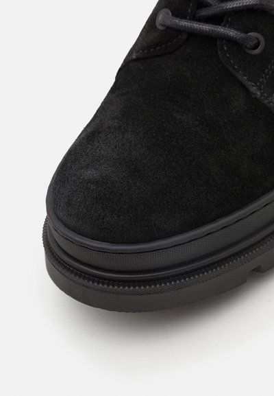 Chaussure en suede noir avec un bout arrondi, une surface texturée et une semelle en caoutchouc noir avec des détails striés. Design simple à lacets.