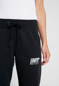 Svarta bomullsbyxor i sweatpants-stil har en ribbad midja med dragsko och en vit logotyp på vänster ben. Slät yta, slim fit-design.