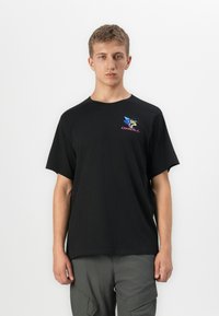Joven vistiendo una camiseta negra de O'Neill con un logotipo colorido en el pecho y pantalones cargo grises, de pie contra un fondo blanco.