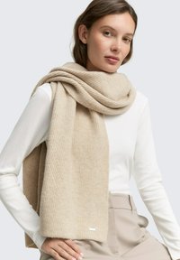 Écharpe beige côtelée en matériau doux, drapée librement autour du cou, présentant une texture subtile et un petit accent de logo.
