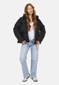 Giubbotto trapuntato nero con zip frontale, cappuccio e lacci regolabili, indossato sopra una maglietta bianca e abbinato a jeans azzurri chiari.