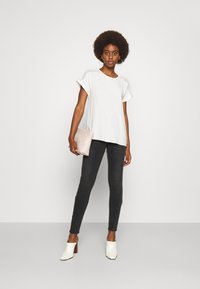 Top branco de manga curta com detalhes texturizados nos ombros, combinado com jeans skinny pretos e sapatos de salto abertos brancos. Asegurando uma carteira rosa clara.