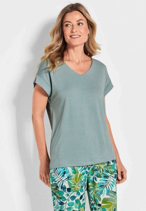 Helles grünes V-Ausschnitt-T-Shirt mit kurzen hochgerollten Ärmeln, aus weichem Stoff. Kombiniert mit bunten Blumenmuster-Hosen mit blauen und grünen Blättern.