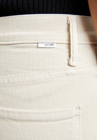 Pantalon en denim crème avec une finition texturée et une étiquette visible portant la mention "MOTHER". Comprend des poches standard et des coutures renforcées.