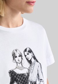 Fehér póló, fekete vázlatokkal ábrázolt két nővel, ékszerekkel díszítve; az egyik mintás öltözetben, a másik csíkos mintázatban.