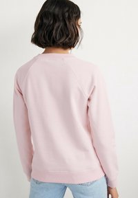 Donna con capelli scuri fino alle spalle, che indossa un felpa a maniche lunghe rosa chiaro e jeans azzurri, di spalle contro uno sfondo neutro.