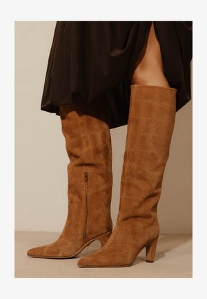 Bottes hauteur genou en suède marron clair avec motif texturé et fermeture éclair latérale, portées avec une jupe noire.