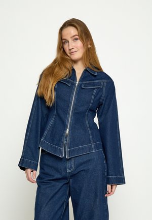 Mladá žena s dlhými červenými vlasmi, oblečená v tmavomodrej rifľovej bunde na zips s bielym prešívaním a zodpovedajúcimi širokými džínsami.