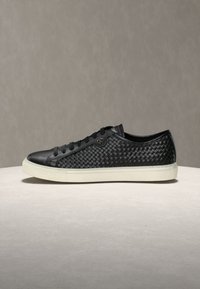 Baskets basses noires avec côtés en cuir tressé, lacets noirs et semelle en caoutchouc blanche sur un fond neutre.