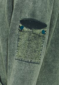 Tissu de sweat-shirt vert olive avec une finition texturée, doté d'une poche plaquée avec des boutons-pression bleus et des détails de coutures contrastantes.