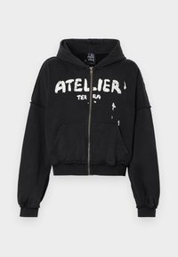 Hoodie preto cropped feito de tecido suave, apresentando um fecho na frente, um grande bolso frontal e a gráfica "ATELIER TERRA" em texto branco.