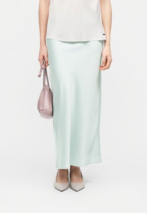 Kobieta ubrana w jasnopistacjową, satynową spódnicę do kostek, białą bluzkę, beżowe szpiczaste szpilki, trzymająca w lewej ręce małą różową torebkę na troczek.