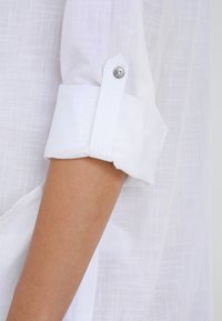 Hvit lin skjorte med oppbrettede ermer, med knappdetalj og teksturert, lett gjennomsiktig stoff som fremhever et lett og luftig design.