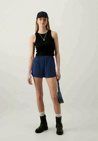 Young woman wearing a black tank top, blue shorts, black cap, black boots, white socks, hoop earrings, necklace, holding small denim handbag.