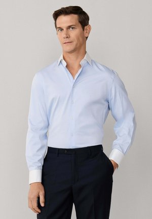 Homme portant une chemise bleu clair à col et poignets blancs, ainsi qu'un pantalon foncé, debout avec une main dans la poche devant un fond uni.