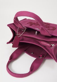 Petit sac à main en tissu violet avec deux poignées, compartiment zippé ouvert, bouton-pression et une étiquette rectangulaire suspendue à une chaîne.