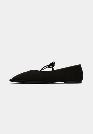 Scarpa piatta nera in suede con punta affilata e sottile laccio annodato a fiocco sulla parte superiore del piede, mostrata di lato su uno sfondo bianco.
