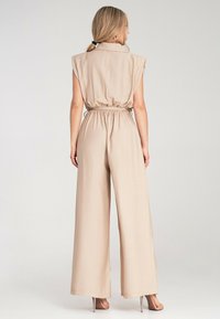 Beżowy kombinezon z detalem w postaci paska, szerokimi nogawkami i krótkimi rękawami. Wykonany z miękkiego materiału, z kołnierzykiem i minimalnymi szwami.