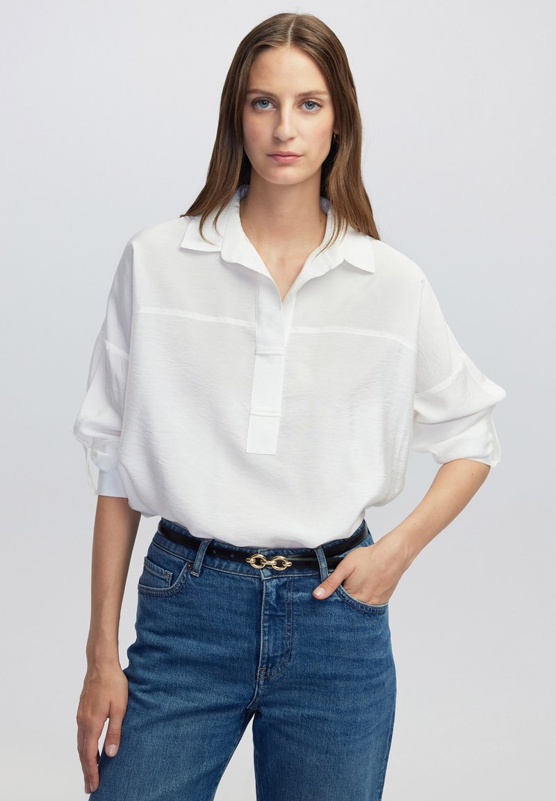 Witte, lichte blouse met een kraag en een knoopsluiting. Lange mouwen opgerold, gecombineerd met hoge blauwe jeans en een zwarte riem.