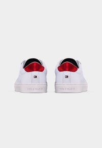 Paire de baskets blanches avec des accents métalliques rouges au talon et de petits logos rectangulaires au-dessus de la semelle embossés avec "Hilfiger" à l’arrière.