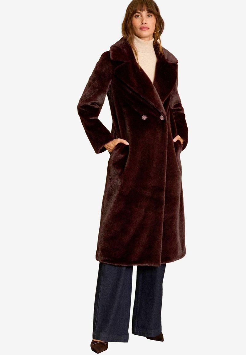 Cappotto in pelliccia finta color bordeaux con design doppiopetto, grande colletto e tasche laterali; indossato sopra pantaloni larghi scuri.