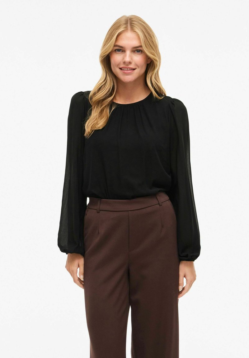 Blusa nera a maniche lunghe con scollatura arricciata e maniche a palloncino trasparenti, abbinata a pantaloni marroni a vita alta. Tessuto morbido e fluido.