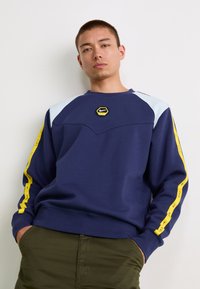 Námornícky sveter s svetlomodrými ramennými akcentami, okrúhlym výstrihom, hexagonálnym žltým logom Nike a žltými bočnými pruhmi s textom značky.