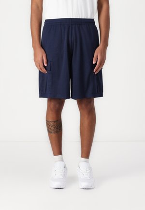 Navy mesh athletic shorts feature an elastic waistband, two side pockets, and a loose fit. Worn with white sneakers and ankle socks.