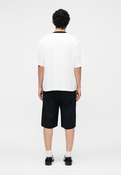 Άνδρας με σγουρά μαλλιά που φοράει λευκό πουκάμισο με κοντά μανίκια, μαύρα σορτς μέχρι το γόνατο, λευκές κάλτσες και μαύρα παπούτσια, στέκεται με την πλάτη προς τα εμπρός.