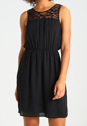 Vestido negro sin mangas con cintura elástica y detalles de encaje en la parte superior del pecho y los hombros, usado por una persona contra un fondo liso.