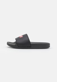 Black slide sandals with a textured strap and a red logo. Soft material, flat sole, and minimalistic design suitable for casual wear.