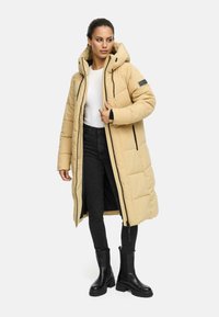 Long, padded coat in light beige with a zip closure and hood. Features black accents and a textured finish. Paired with black jeans and boots.
