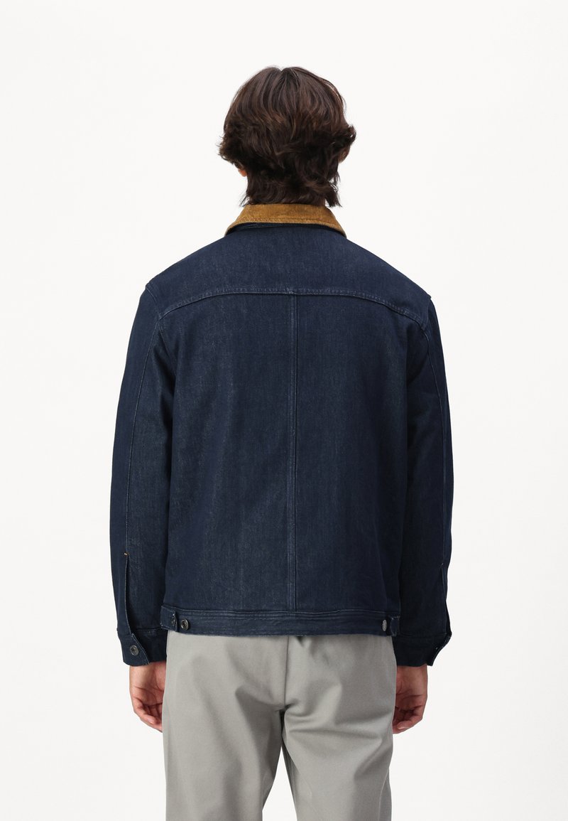 Jeansowa kurtka w ciemnym niebieskim kolorze, z brązowym kołnierzem z sztruksu, zapięciem na guziki przy mankietach i prostym krojem. Z tyłu widoczne szczegóły szycia.