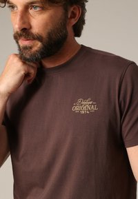 T-shirt en coton brun à col rond, avec un logo doré imprimé qui indique "Deluxe Original 1974" sur la partie gauche de la poitrine.
