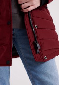 Mørkerød pufferjakke med et tekstureret, quiltet design, der har en lynlåslukning og trykknapper i bunden. Parret med jeans.