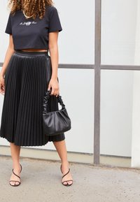 T-shirt noir court avec texte brodé, jupe midi plissée noire, sandales noires à lanières, et un petit sac à main noir avec une poignée froncée.