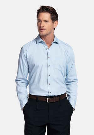 Man draagt lichtblauw overhemd met knopen, donkere broek, bruine riem, handen in zakken, kijkt opzij tegen een effen achtergrond.